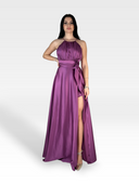 Una donna indossa con sicurezza un abito di raso lungo fino al pavimento, senza maniche, con un alto spacco sul lato. L'abito 18teen VIOLA presenta uno scollo all'americana e una cintura annodata che ne accentua la vita. Posa con una mano sul fianco e l'altra appoggiata accanto a lei, mantenendo un'espressione neutra.