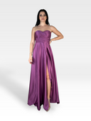 Una donna indossa con sicurezza un abito di raso magenta senza spalline, lungo fino al pavimento, con scollatura a cuore e uno spacco alto. Indossa tacchi alti argentati e ha i capelli lunghi e scuri. Lo sfondo è un semplice bianco sfumato. Indossa l'abito LAYTON di 18teen.