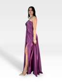 Una donna che indossa un LAYTON di 18teen viola senza spalline, in raso, lungo fino al pavimento, con uno spacco alto fino alla coscia e scollo a cuore, si staglia su un semplice sfondo bianco. Ha lunghi capelli neri e indossa tacchi alti argentati e un braccialetto al polso sinistro. Si guarda alle spalle, sorride leggermente nel suo abito elegante.