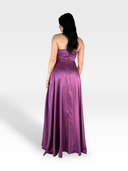 Una persona con lunghi capelli scuri è in piedi con le spalle alla telecamera, indossando un LAYTON 18enne, un abito di raso viola senza spalline e lungo fino al pavimento. L'abito vanta un corpetto aderente con scollo a cuore che sfocia in un'ampia gonna su un semplice sfondo bianco sfumato.