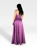 Una persona con capelli lunghi e scuri sta di fronte, mostrando la parte posteriore di un ALYSSUM di raso viola setoso lungo fino al pavimento di 18teen con spalline sottili. Lo sfondo è bianco semplice, sottolineando la silhouette fluida dell'abito e il tessuto lucido.