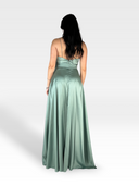 Una donna con lunghi capelli scuri è rivolta lontano dalla telecamera, mostrando la parte posteriore di ELVENE di 18teen, il suo vestito verde fluente di raso. L'abito ha un corpetto aderente con una piccola fila di bottoni e maniche con cinturino, che conduce a un'elegante gonna lunga. L'abito in raso contrasta magnificamente con il semplice sfondo bianco.