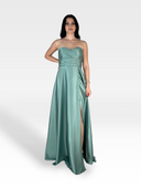Una donna con lunghi capelli scuri indossa un abito di raso verde acqua, senza spalline, lungo fino al pavimento. L'AZUREA di 18teen presenta uno scollo a cuore e uno spacco alto sulla gamba, mettendo in mostra il suo design raffinato. Si staglia con sicurezza su uno sfondo semplice, grigio chiaro, trasudando un'eleganza sofisticata.