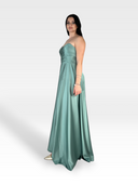 Una donna con un lungo abito verde acqua senza spalline si staglia su uno sfondo sfumato grigio chiaro. Ha i capelli scuri e indossa braccialetti su un braccio. Trasuda eleganza sofisticata l'AZUREA di 18teen, con tacchi alti dorati appena visibili sotto il vestito.