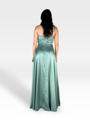 Una donna con lunghi capelli scuri sta dando le spalle alla telecamera, indossando un abito senza spalline in raso AZUREA, lungo fino al pavimento, del 18esimo secolo, in un colore verde pallido. L'abito presenta un corpetto increspato e una gonna fluida, che incarna l'eleganza sofisticata. Lo sfondo è semplice e grigio chiaro.