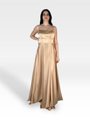 Una donna con lunghi capelli scuri indossa un IMMAN lungo fino al pavimento e senza maniche in raso color champagne. In piedi su uno sfondo semplice, grigio chiaro, con una mano sul fianco e un'espressione neutra, trasuda eleganza senza tempo.