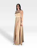 Una donna con lunghi capelli scuri indossa un abito IMMAN in raso color champagne lungo fino al pavimento di 18teen con spalline sottili, che irradia eleganza senza tempo. Lei sta in una posizione neutra su un semplice sfondo bianco, accessoriata con braccialetti e ballerine dorate.