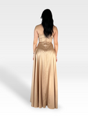 Una persona con lunghi capelli scuri è in piedi e guarda dall'altra parte, indossando un abito LAKME by 18teen in raso, senza spalline, lungo fino al pavimento in una tonalità color champagne. L'abito presenta un corpetto aderente con una fila di bottoni sul retro e una gonna fluida. Lo sfondo è una sfumatura semplice e leggera, che incarna l'eleganza senza tempo.